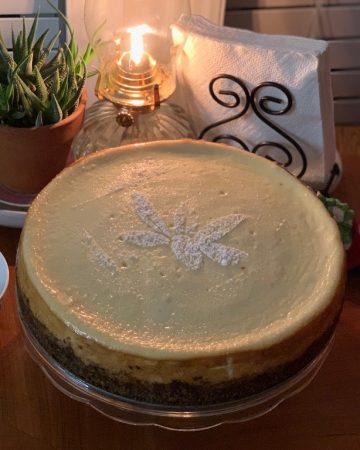 Classic New York Style Cheesecake topped with stenciled powdered sugar poinsettia flower, displayed on dining table with strawberry and chocolate ganache sauces on side.