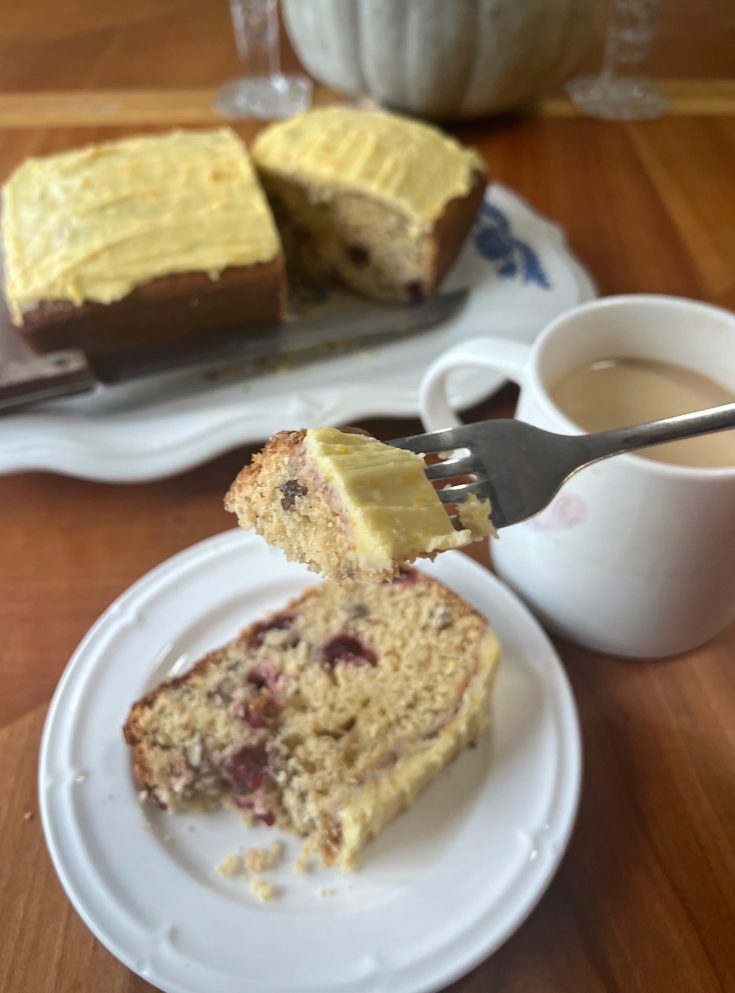 Bite of cranberry orange sourdough loaf on a fork beside a plated slice, coffee, and the frosted loaf.