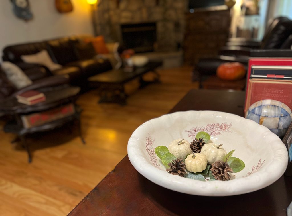 A vintage floral bowl holding small cream pumpkins, pinecones and greenery beside old books