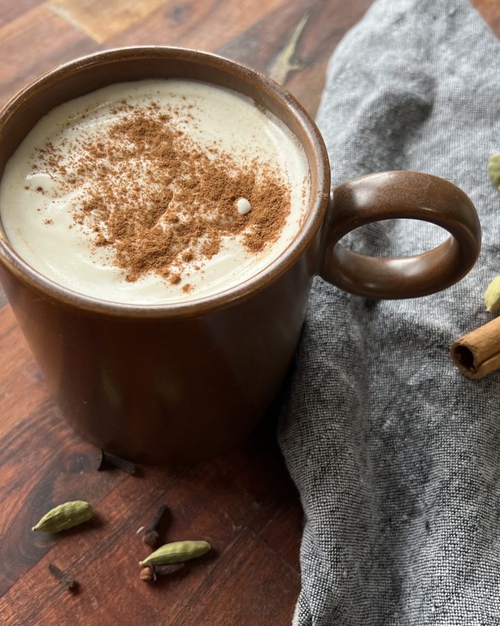 Pumpkin chai tea latte topped with maple whipped cream and cinnamon in a brown mug beside whole spices.