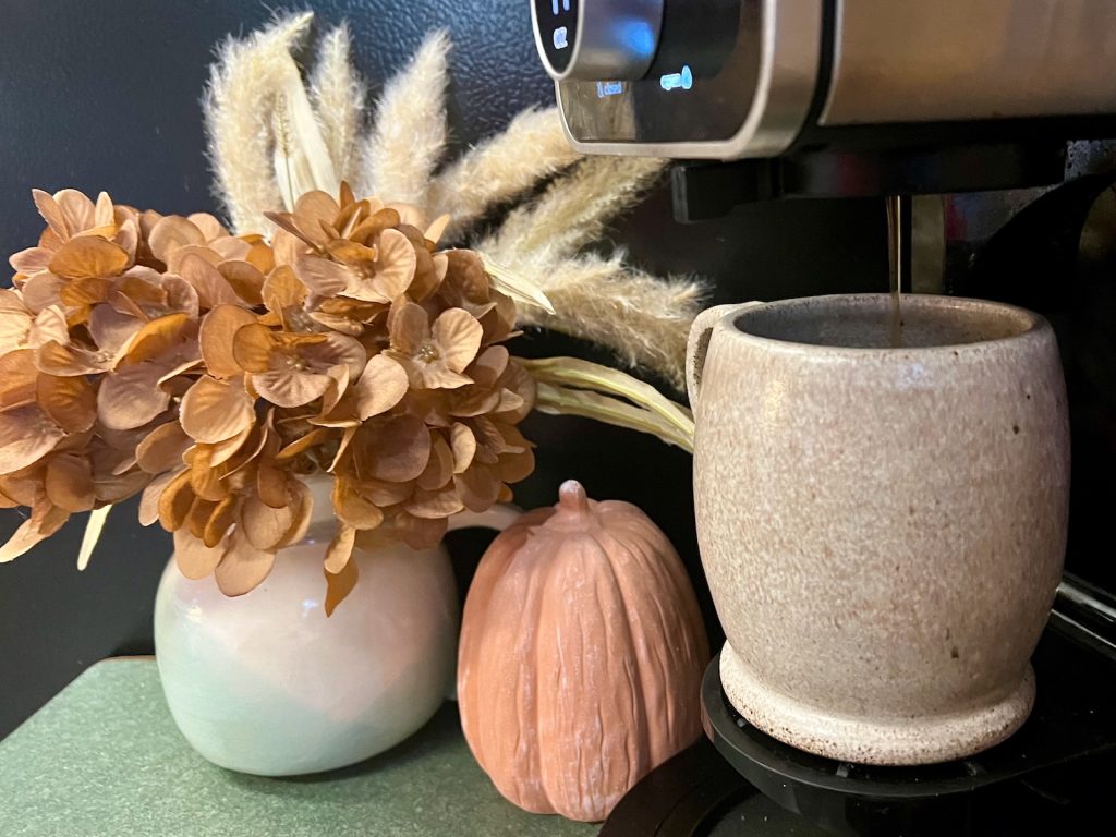 Coffee brewing into a stoneware mug beside dried hydrangeas and a small terracotta-colored pumpkin