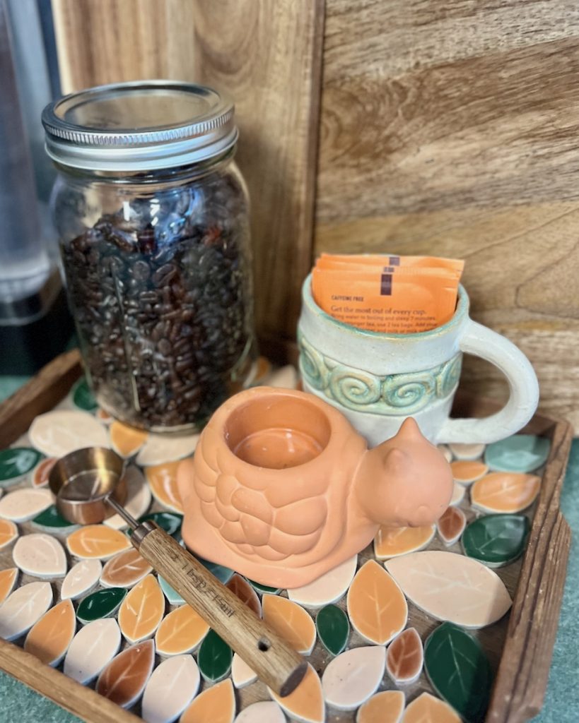 Coffee beans in a mason jar with tea packets and a wooden-handled scoop on a patterned tray.