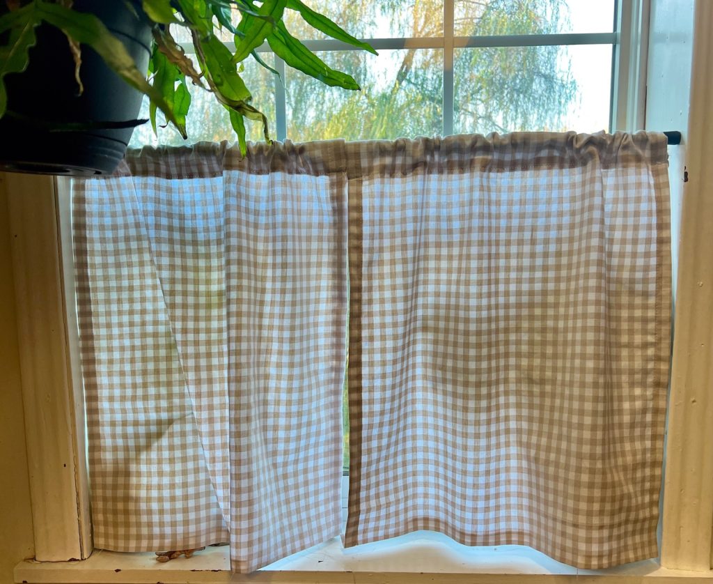 Two finished hand sewn tan gingham café curtains hung in the window on a brown tension rod.
