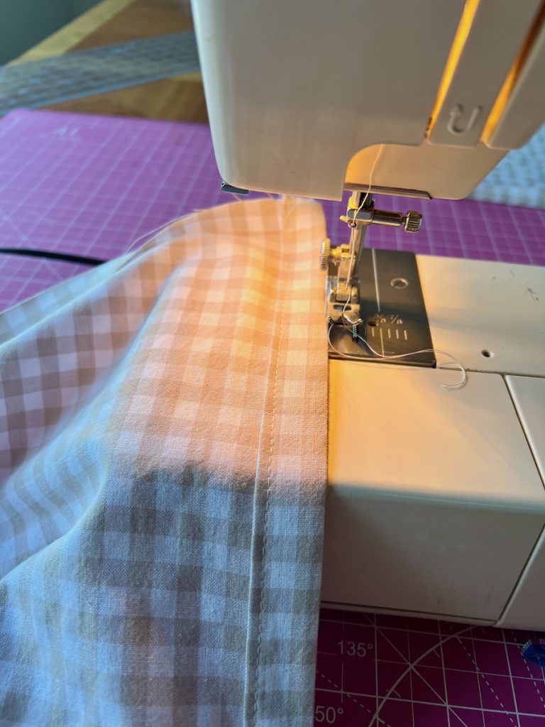 Close up of finished side hem of DIY gingham café curtains resting on a sewing machine next to the needle and thread.