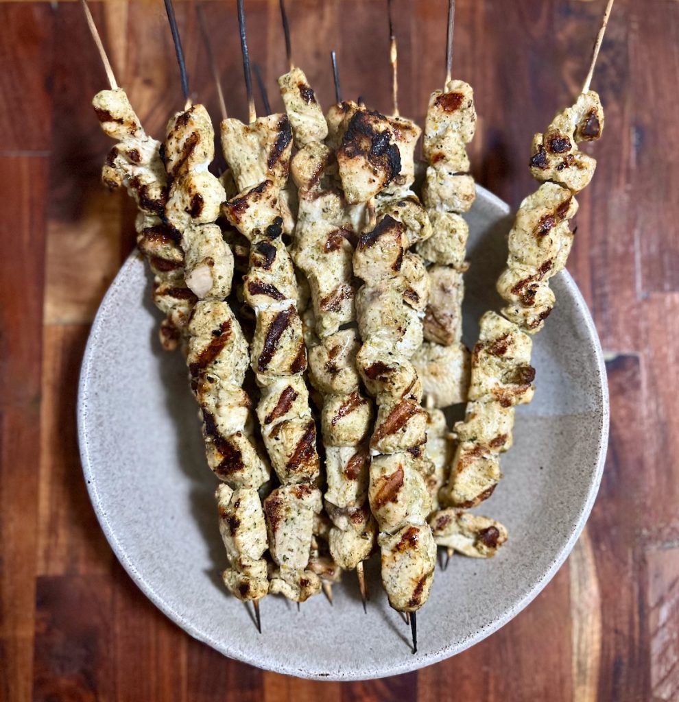 Grilled chicken skewers with browned edges and herbs on a gray serving platter.