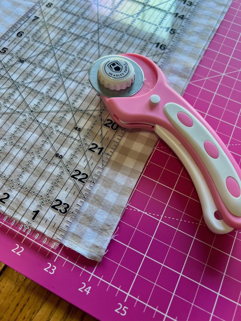 Rotary cutter and quilting ruler used to trim beige gingham fabric for café curtains.