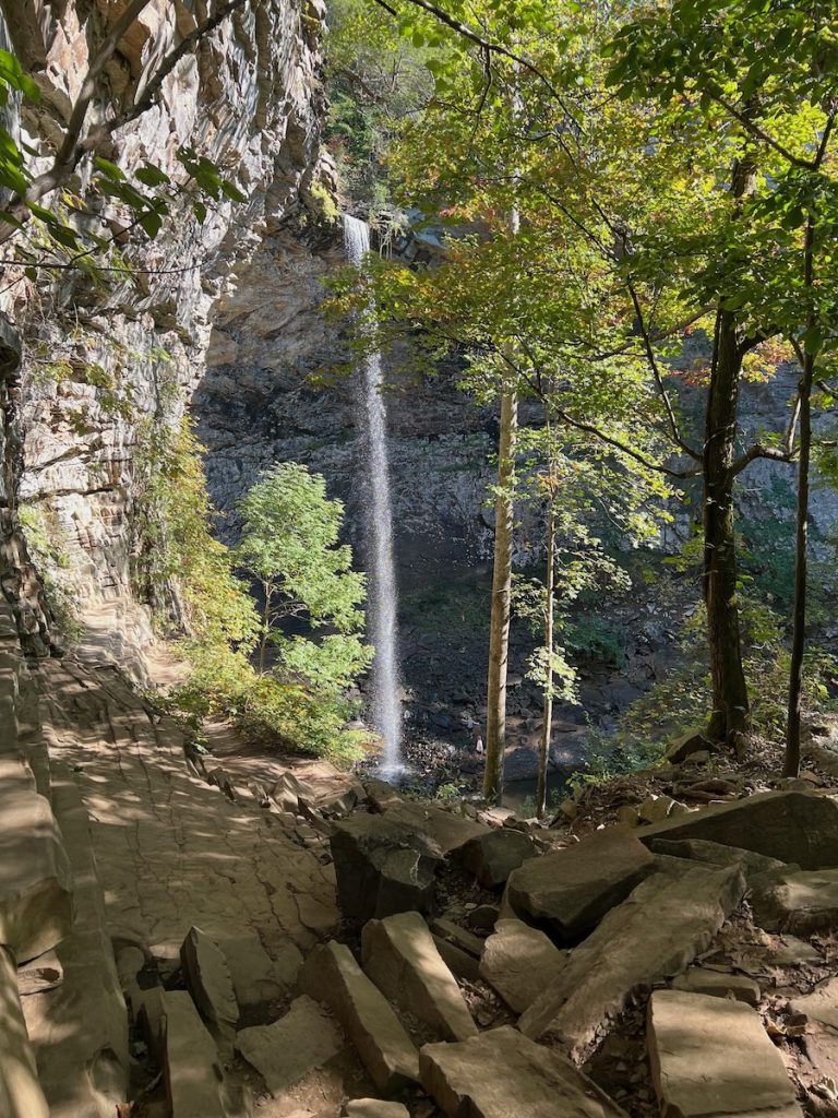 Tall waterfall dropping over a rocky cliff, framed by leafy trees and large boulders.